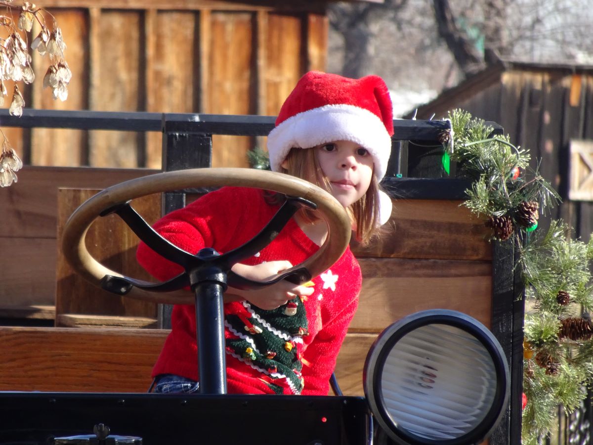 Child wearing Santa hat and a sweatshirt with a Christmas tree on it sits at the wheel of an antique vehicle that is decorated with an evergreen wreath.