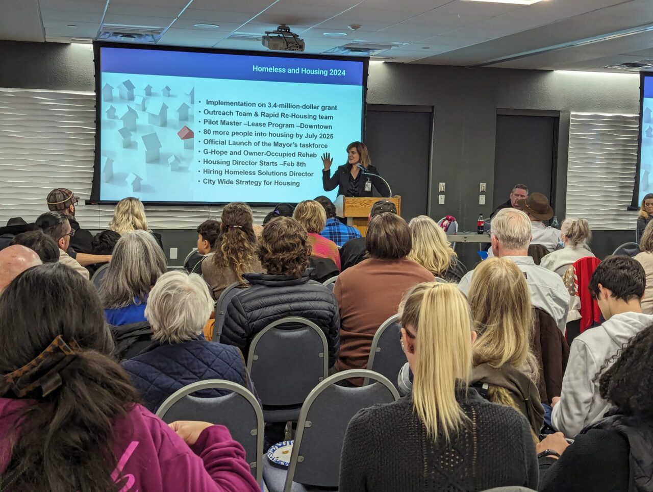 A woman stands behind a podium and speaks to a crowd of people. Behind her is a screen with a list of homeless and housing projects and initiatives that are coming in 2024 and 2025.