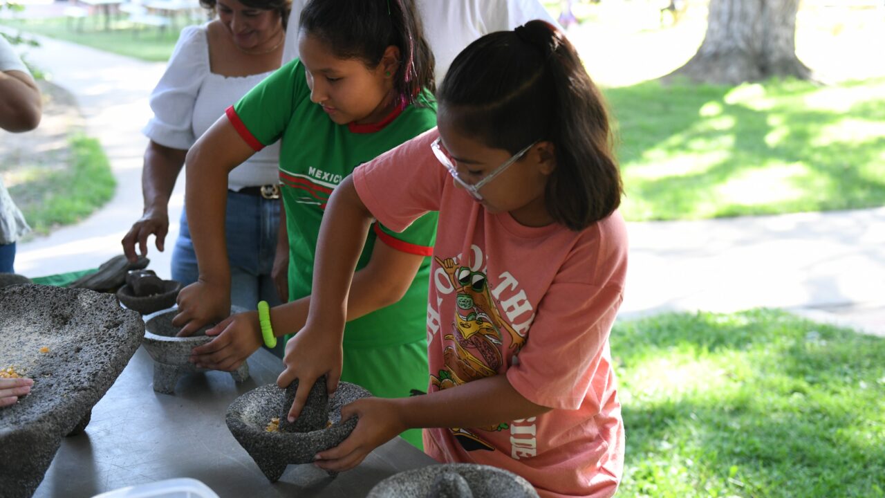Children learn to grind corn by hand on a metate, a bowl made from stone.