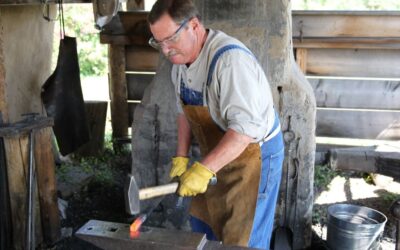 A man with heavy gloves and a blacksmithing apron works at a forge to hammer a piece of hot metal into a shape.