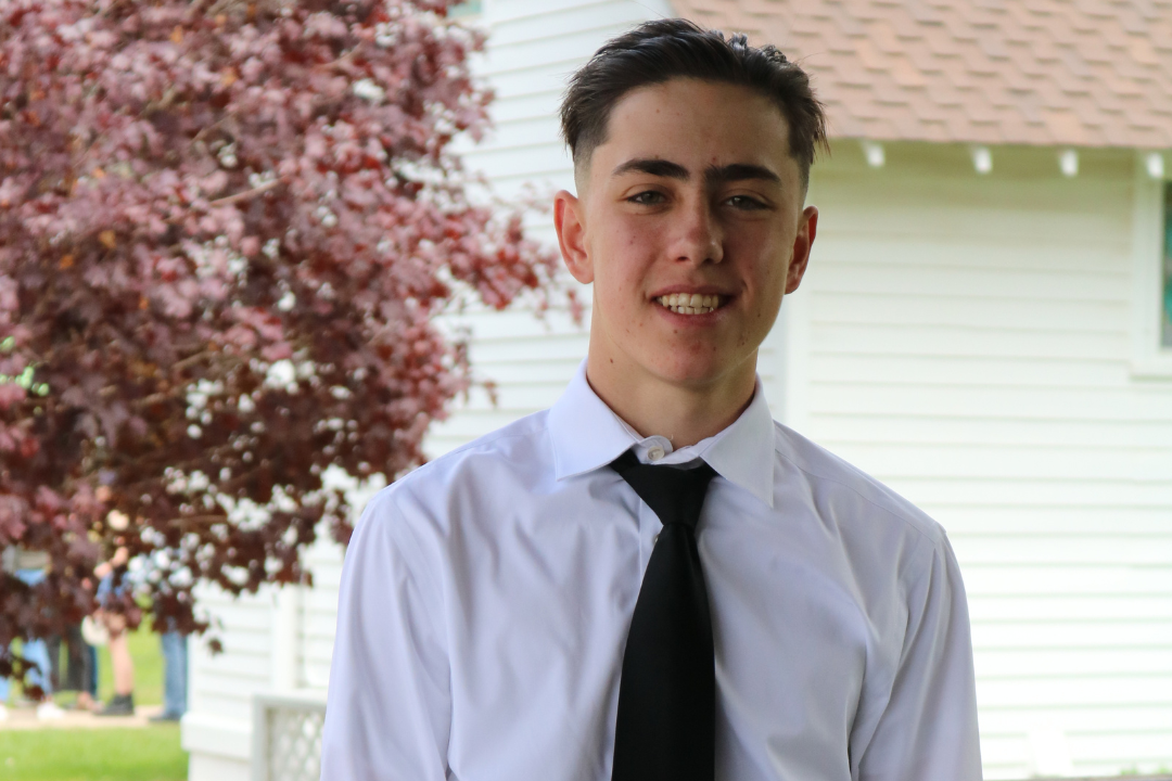 A young man dressed in a collared shirt, tie and pants stands in front of a railing with a slight smile. There is a tree and a house in the background.