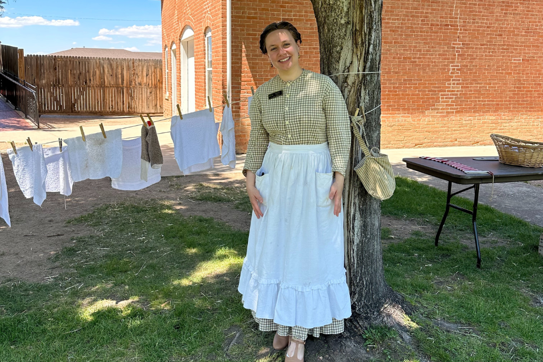 Katie Henry, costumed in a gingham dress and white cotton apron, stands near a clothing line and brick building at Greeley's historical Centennial Village Museum.