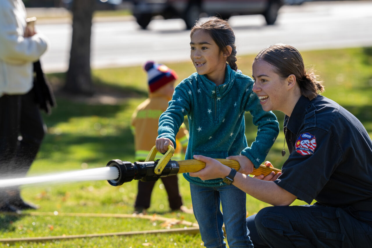 Female firefighter helping a young girl use a fire hose at an event, with a young child dressed in a firefighter outfit standing in the background.