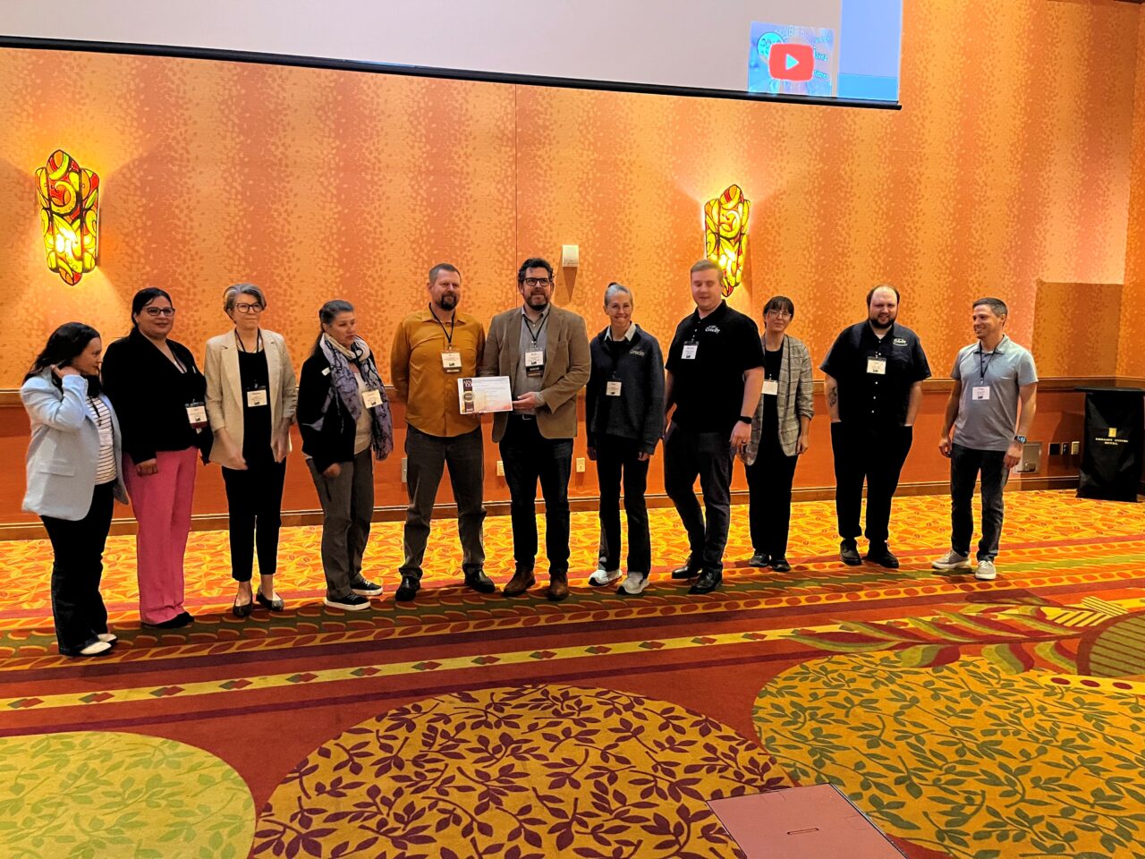 A group of eleven people indoors with one person holding a certificate. The group is dressed in business casual attire, standing along a decorated wall in a warmly lit room.