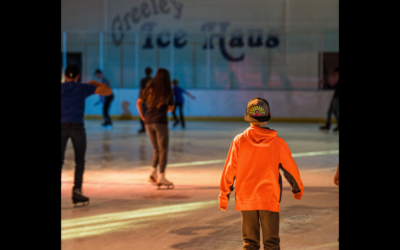 A young child in an orange shirt and backward hat skates towards others on the brightly colored Greeley Ice Haus rink.