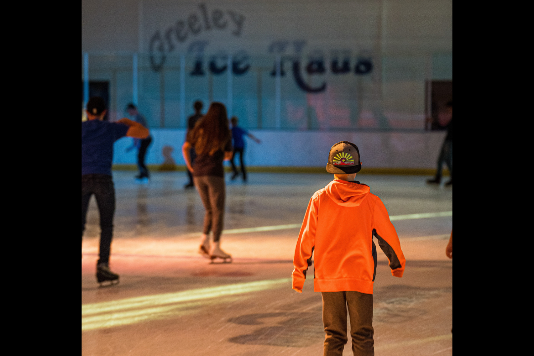 A young child in an orange shirt and backward hat skates towards others on the brightly colored Greeley Ice Haus rink.