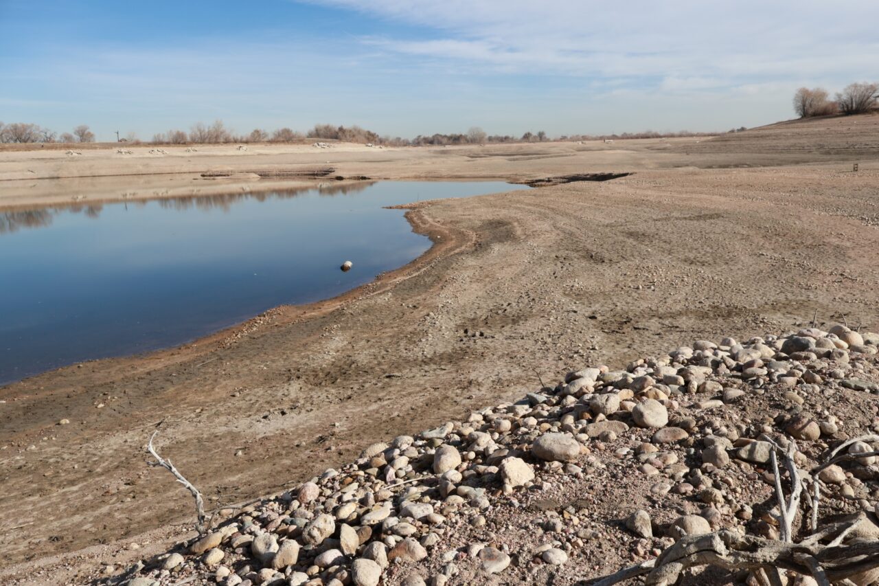 A dry, barren pond with a small patch of water remaining near the center, surrounded by sandy, rocky shores.