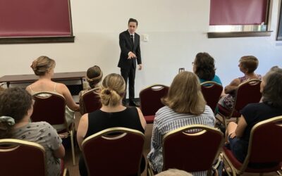 A young person wearing a black suit presents to a crowd of people seated in the community room at the Greeley History Museum.