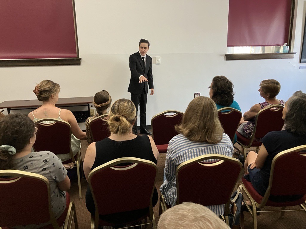 A young person wearing a black suit presents to a crowd of people seated in the community room at the Greeley History Museum.