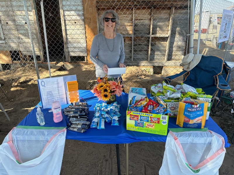 Susan Cribelli stands behind a table covered with snacks, a bouquet of flowers and trash bags at the Dearfield Day event. The former Dearfield filling station is in the background.