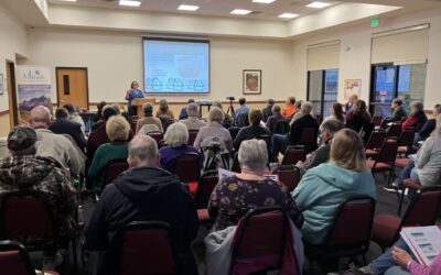 A person stands behind a podium giving a presentation in front of a room full of people seated in chairs. There is a screen on the wall with a PowerPoint presentation shown.
