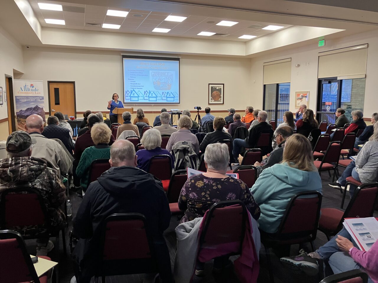 A person stands behind a podium giving a presentation in front of a room full of people seated in chairs. There is a screen on the wall with a PowerPoint presentation shown.