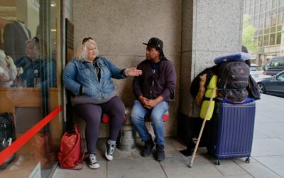 A man and a woman seated on chairs on a downtown sidewalk in front of a large building. They appear to be talking. The woman is pointing towards the street. Luggage and belongings are nearby.
