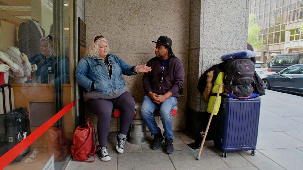 A man and a woman seated on chairs on a downtown sidewalk in front of a large building. They appear to be talking. The woman is pointing towards the street. Luggage and belongings are nearby.