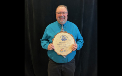 A smiling man wearing glasses and a button-down shirt stands in front of a black backdrop and holds a round wooden plaque engraved with “Columbine Award for Renovated Park Design” and the Colorado Parks and Recreation Association logo.