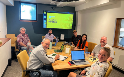 gBETA Greeley cohort participants and mentors smile for a photo around a conference table during a “Revenue Models” session. A presentation slide is visible on the screen at the back of the room.