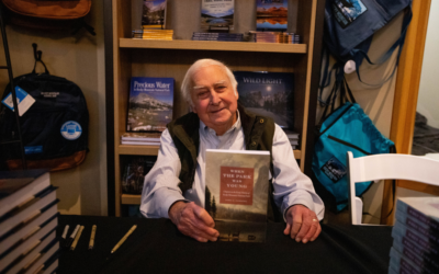 A man holds a book he wrote, surrounded by a bookshelf.