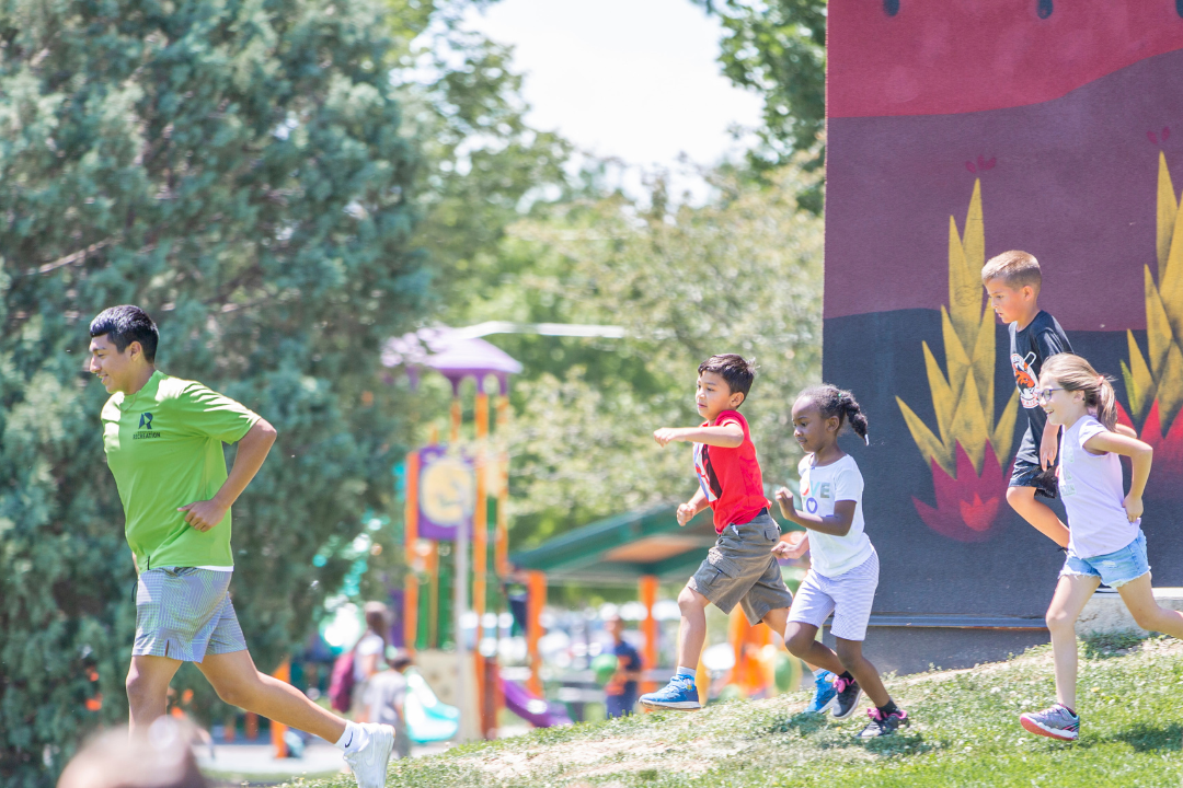 A recreation staff member leads a group of young children running down a grassy hill at Hoshiko Park on a sunny day. The kids are smiling and active, with a colorful playground and mural out of focus in the background.
