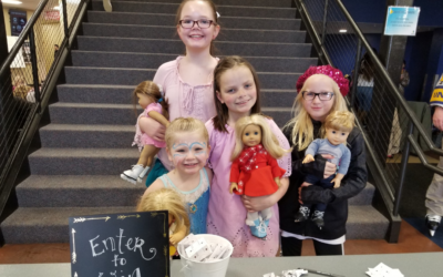 Four girls smile holding their American Girl Dolls by a raffle cup that reads "enter to win" at the Greeley Ice Haus.