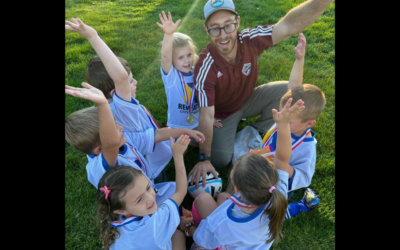 Volunteer soccer coach holds ball and poses with team in a circle on a grassy field.
